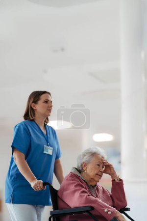 Foto de Enfermera empujando a un paciente mayor en una silla de ruedas a través del pasillo del hospital, hall. Apoyo emocional para la mujer anciana. - Imagen libre de derechos