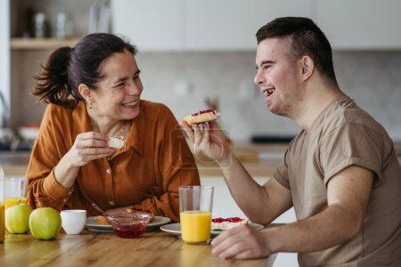 Photo for Young man with Down syndrome having breakfast with his mother at home. Morning routine for man with Down syndrome genetic disorder. - Royalty Free Image