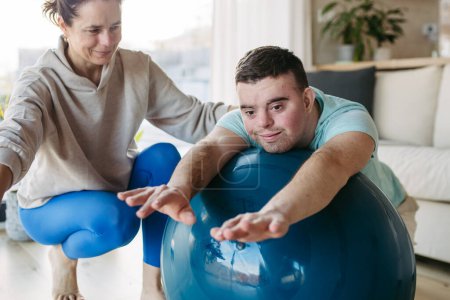Foto de Hombre joven con síndrome de Down haciendo ejercicio en casa con su madre en una pelota de fitness. Rutina de entrenamiento para discapacitados. - Imagen libre de derechos