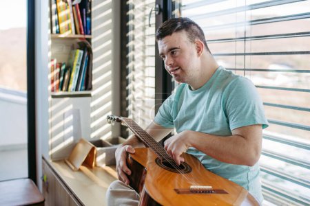 Young man with down syndrome playing acoustic guitar, sitting by window, holding and strumming a guitar, making beautiful music.