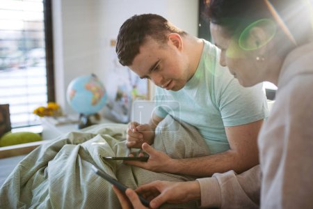 Young man with down syndrome sitting in his bed with mom, both looking at smartphone in the morning. Morning routine for man with disability.