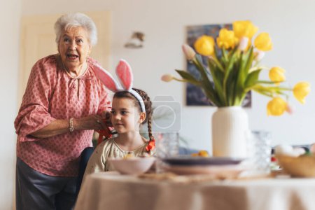 Großmutter flechtet Enkelinnen Haare, legt rote Schleifen an Zöpfe. Vorbereitung auf das Osteressen.. Konzept der Osterferien für Familien.