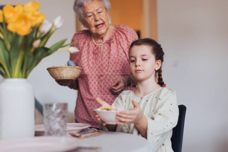 Photo for Grandmother with granddaughter setting table for traditional easter lunch. Recreating family traditions and customs. Concept of family easter holidays. Standing by table, praying. - Royalty Free Image