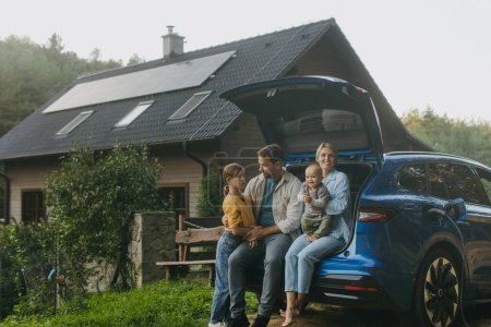 Foto de Familia con coche eléctrico de pie en frente de su casa con paneles solares en el techo. Energía solar y estilo de vida sostenible de la familia joven. Concepto de energía verde y futuro sostenible para el próximo - Imagen libre de derechos