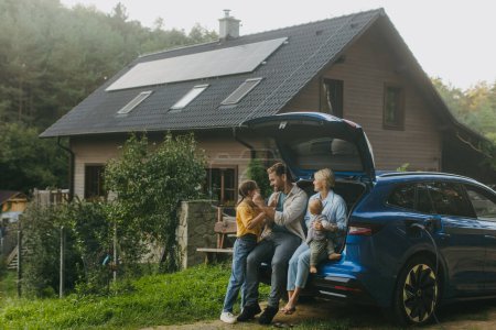 Téléchargez les photos : Famille avec voiture électrique debout devant leur maison avec des panneaux solaires sur le toit. Énergie solaire et mode de vie durable de la jeune famille. Concept d'énergie verte et d'avenir durable pour la prochaine - en image libre de droit