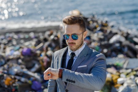Fashionable modern businessman looking at watch, checking time standing on pile of waste on beach. Consumerism versus pollution concept. Corporate social responsiblity in business.