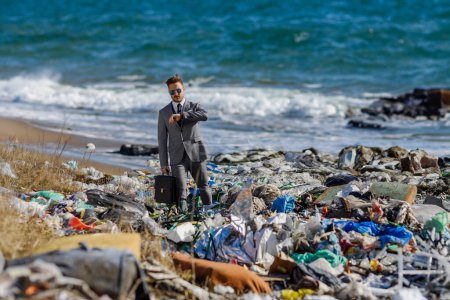Foto de Moderno hombre de negocios de moda mirando el reloj, comprobando el tiempo de pie en la pila de residuos en la playa. Concepto de consumo versus contaminación. Responsabilidad social corporativa en los negocios. - Imagen libre de derechos