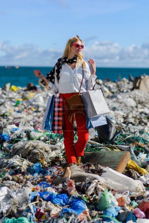 Fashionable modern woman on landfill with shopping bags, standing on pile of waste on beach. Consumerism versus pollution concept.