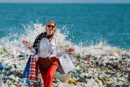 Fashionable modern woman on landfill with shopping bags, standing on pile of waste on beach. Consumerism versus pollution concept.