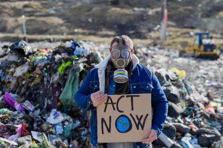 Téléchargez les photos : Homme militant avec masque à gaz portant une pancarte, signe de protestation, debout sur la décharge, grand tas de déchets, concept environnemental et éco-activisme. - en image libre de droit