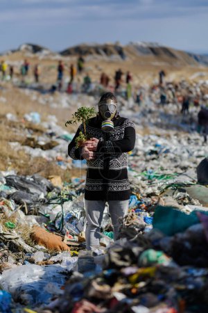 Téléchargez les photos : Homme avec masque à gaz tenant une usine en pot sur une décharge debout sur une décharge, grand tas de déchets, concept environnemental et éco-activisme. - en image libre de droit