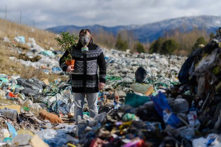 Foto de Hombre con máscara de gas sosteniendo planta en maceta en vertedero de pie en el vertedero, gran pila de residuos, concepto ambiental y activismo ecológico. - Imagen libre de derechos