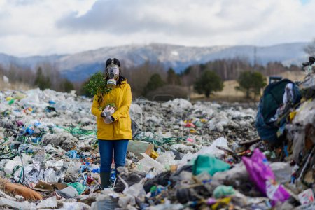 Foto de Activista femenina con máscara de gas sosteniendo planta en maceta sobre vertedero, gran pila de residuos, concepto ambiental y activismo ecológico. - Imagen libre de derechos