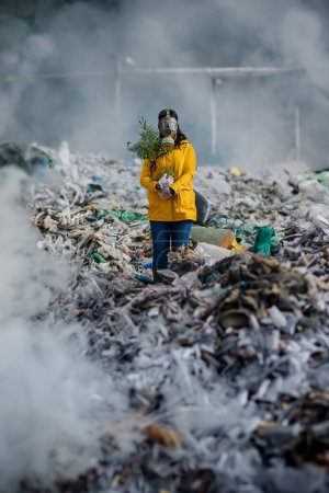 Téléchargez les photos : Militante avec masque à gaz tenant une plante en pot debout sur une décharge, une grande pile de déchets, un concept environnemental et un éco-activisme. - en image libre de droit