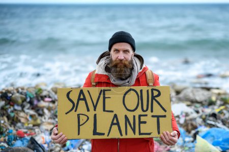Male activist holding placard, protest sign, standing on landfill, large pile of waste on sea beach, shoreline, environmental concept and eco activism.