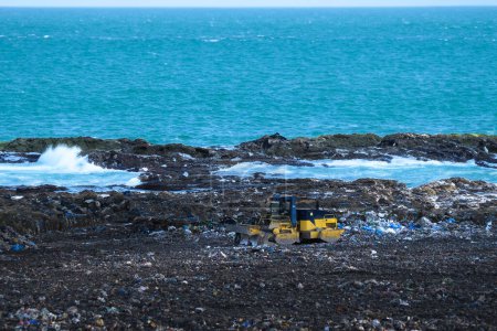Garbage excavator pushing a pile of waste on coast, sea shoreline. Marine debris, accumulation on beaches, global problem, environmental concept. Copy space.