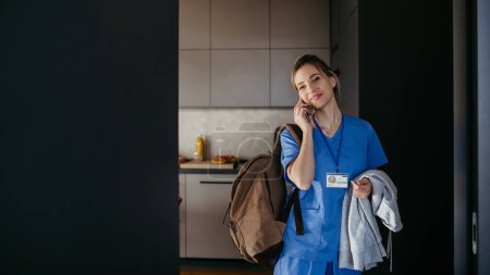 Foto de Enfermera, doctora preparándose para el trabajo, llamando por teléfono a un smartphone, saliendo de casa en bata con mochila. Equilibrio entre vida laboral y personal sanitario. - Imagen libre de derechos