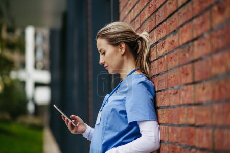 Foto de Enfermera, doctor en uniforme azul tomando descanso, apoyándose en el edificio del hospital, desplazándose en el teléfono inteligente. Equilibrio entre vida laboral y personal sanitario. - Imagen libre de derechos