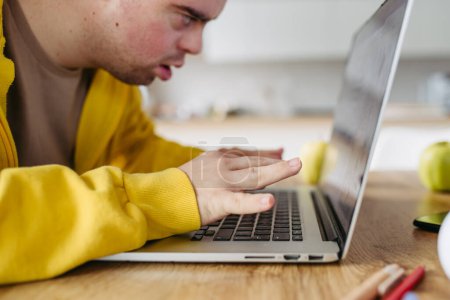 Foto de Hombre joven con síndrome de Down aprendiendo en línea en casa, utilizando el ordenador portátil, trabajando en el portátil. Consulta de telesalud con médico. - Imagen libre de derechos