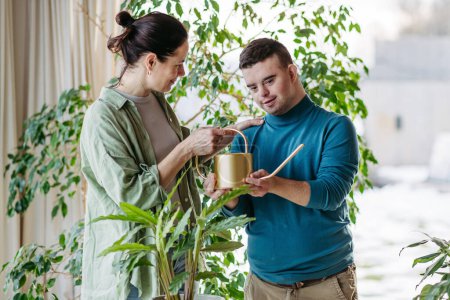 Portrait of young man with Down syndrome with his mother at home, watering houseplant, taking care of plants. Concept of love and parenting disabled child.