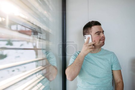 Téléchargez les photos : Portrait de jeune homme trisomique assis par la fenêtre, faisant téléphone avec smartphone. - en image libre de droit