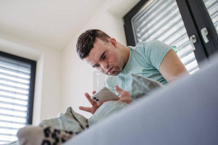 Young man with down syndrome sitting in his bed, looking at smartphone in the morning. Morning routine for man with disability.