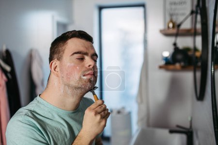Foto de Hombre joven con síndrome de Down aprendiendo a afeitarse, sosteniendo la navaja de afeitar en la mano, mirando al espejo. Vista lateral del hombre discapacitado enfocado. - Imagen libre de derechos