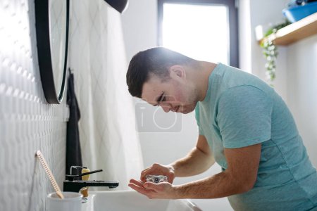 Photo for Young man with down syndrome learning how to shave,applying after shave lotion or cologne on face. Side view of focused disabled man. - Royalty Free Image