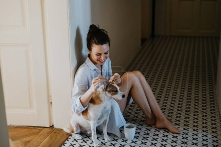 Photo for Woman in pajamas sitting on floor, playing with her cute dog, drinking cup of morning coffee. Weekend morning relaxation at home. Hygge lifestyle. - Royalty Free Image