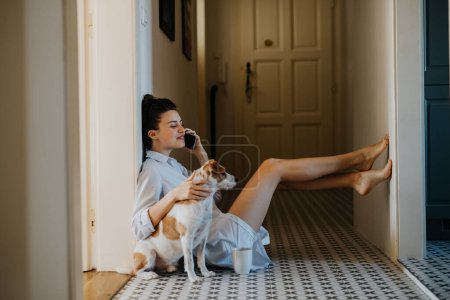 Téléchargez les photos : Femme en pyjama assise sur le sol, téléphone, caressant son chien mignon. Week-end matin détente à la maison, café du matin. Mode de vie Hygge. - en image libre de droit