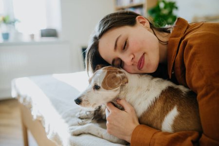 Beautiful single woman lying on bed, snuggling and petting her dog with closed eyes, enjoying weekend. Young woman living alone in apartment with pet,