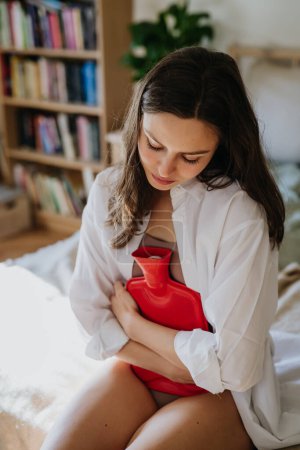 Young woman at home suffering from menstrual pain, having cramps. Woman warming lower abdomen with a hot water bottle, endometriosis, and conditions causing pain in tummy.