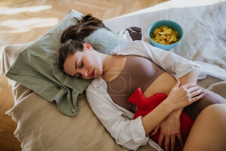 Junge Frau liegt mit Menstruationsschmerzen und Krämpfen im Bett und hat Heißhunger. Frau wärmt Unterbauch mit Wärmflasche, Endometriose und Schmerzen im Bauch.