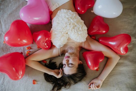 Photo for Sensual woman lying of bed in the middle of heart baloons, dressed in heart shaped crop top. Concept of Valentines Day, love and romantic relationship. - Royalty Free Image