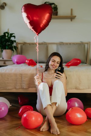 Photo for Beautiful woman sitting by bed in the middle of baloons, scrolling on smartphone. Concept of Valentines Day, love and romantic dating. - Royalty Free Image