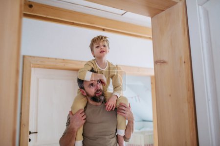 Father carries his son on his shoulders. Boy in pajamas waking up, dad carrying him from bedroom in the morning.