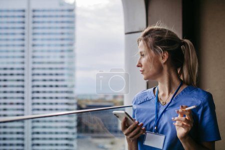 Photo for Nurse smoking cigarette at balcony after work, scrolling on smartphone. Doctor feeling overwhelmed at work, taking break. Stresfull at hospital, clinic. - Royalty Free Image