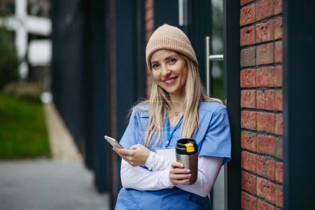 Krankenschwester, Arzt in blauer Uniform macht Pause, lehnt am Krankenhausgebäude, scrollt auf dem Smartphone. Work-Life-Balance im Gesundheitswesen.