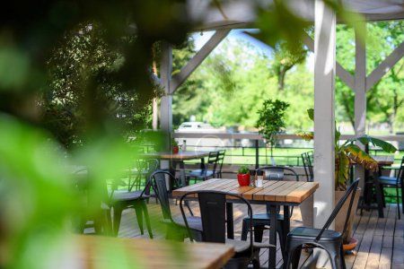 Terraza cubierta de verano de un restaurante con mesas de madera, sillas y suelos. Restaurante patio hecho de materiales naturales con mucha vegetación.