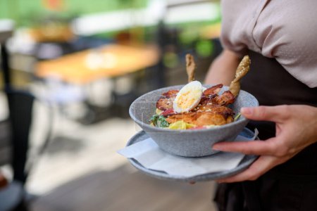 Foto de Cierre de camarera sosteniendo plato con comida, tazón con ensalada de verduras. Servidor en la terraza del restaurante. - Imagen libre de derechos