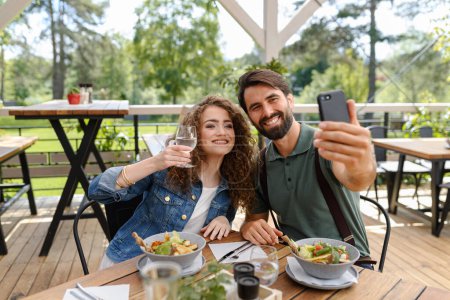 Photo for Young couple at date in restaurant, taking selfie, sitting on restaurant terrace. Boyfriend and girlfriend enjoying springtime, having lunch or brunch outdoors, outdoor seating for dining. - Royalty Free Image