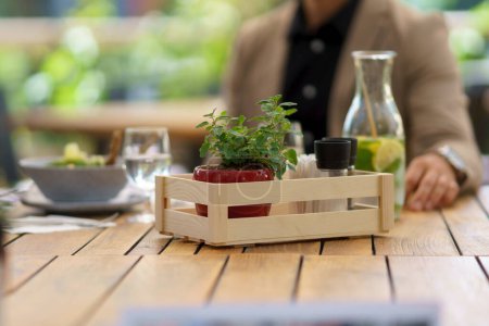 Photo for Wooden table on restaurant patio. Dining table with salt, spices, seasoning containers in crate, small plant in pot as decoration. - Royalty Free Image
