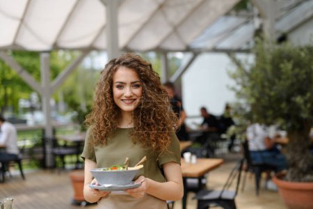 Foto de Retrato de una hermosa camarera sosteniendo el plato con comida, tazón con ensalada. servidor femenino de pie en la terraza del restaurante en un delantal, mirando a la cámara, sonriendo. - Imagen libre de derechos