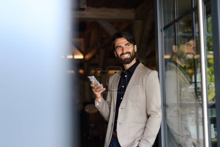 Téléchargez les photos : Portrait d'homme d'affaires mature veste en costume debout à l'intérieur dans un bureau moderne, tenant smartphone dans la main. - en image libre de droit