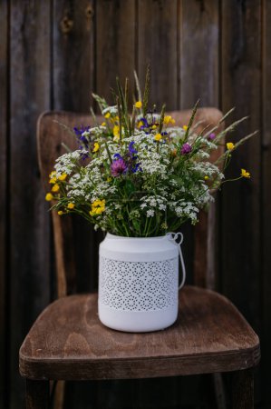 White vase full of meadow flowers, herbs and grass, placed on a wooden chair outdoor. A colorful variety of summer wildflowers.