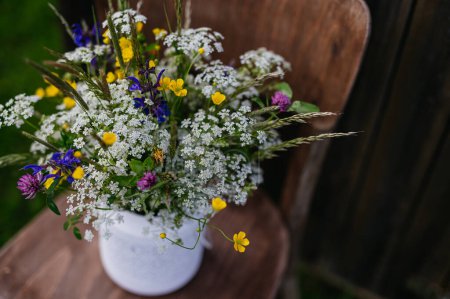 Téléchargez les photos : Vase blanc plein de fleurs de prairie, d'herbes et d'herbe, placé sur une chaise en bois en plein air. Une variété colorée de fleurs sauvages d'été. - en image libre de droit