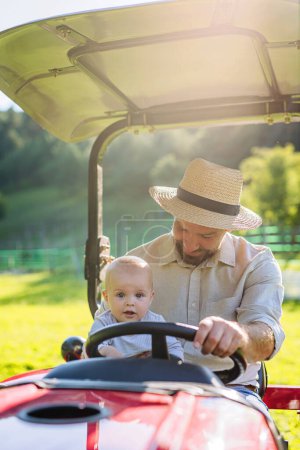 Landwirt Vater auf Traktor mit seinem kleinen Sohn. Kleines Baby wächst auf dem Bauernhof der Familie auf. Konzept der Mehrgenerationenlandwirtschaft.
