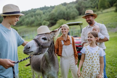 Téléchargez les photos : Famille de fermiers marchant avec l'âne sur leur ferme. Une mule grise portant un chapeau. L'âne comme animal de ferme à la ferme familiale. Concept d'agriculture multigénérationnelle. - en image libre de droit