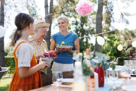 Fille aidant grand-mère et mère à mettre la table pour la fête de jardin d'été. Apporter des assiettes, de la nourriture et des boissons. Les générations d'arbres de la femme en famille.