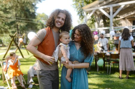Young parents holding baby at a family garden party. Father, mother, and a small child at birthday party.